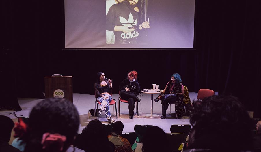 A panel discussion with three people on stage in a dimly lit auditorium at Big Creative Academy in Walthamstow, Waltham Forest. A projected image show