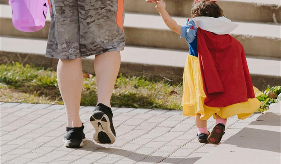 Little girl in costume with with an adult holding a purple pumpkin bucket