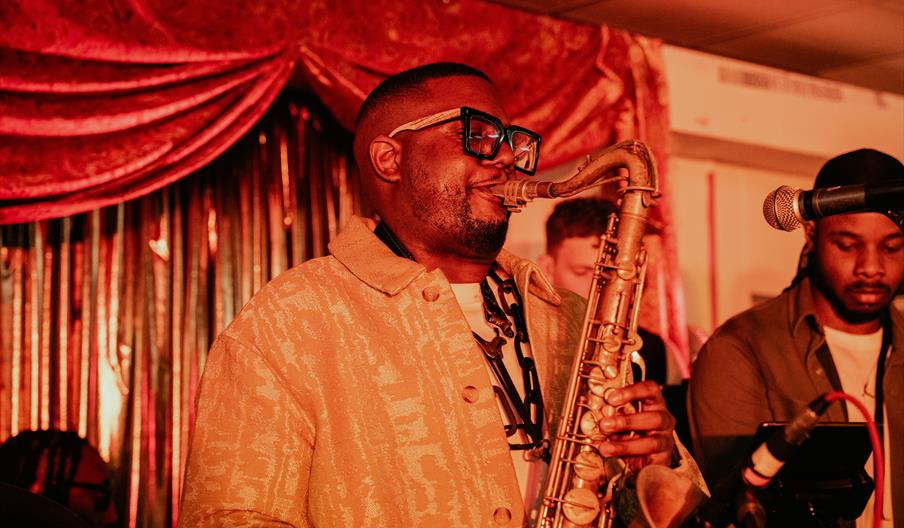 Lewis Daniel, Jazz artist and saxophonist, wearing glasses plays passionately in a cozy, warmly lit venue in Leytonstone. Next to him, a man focuses o